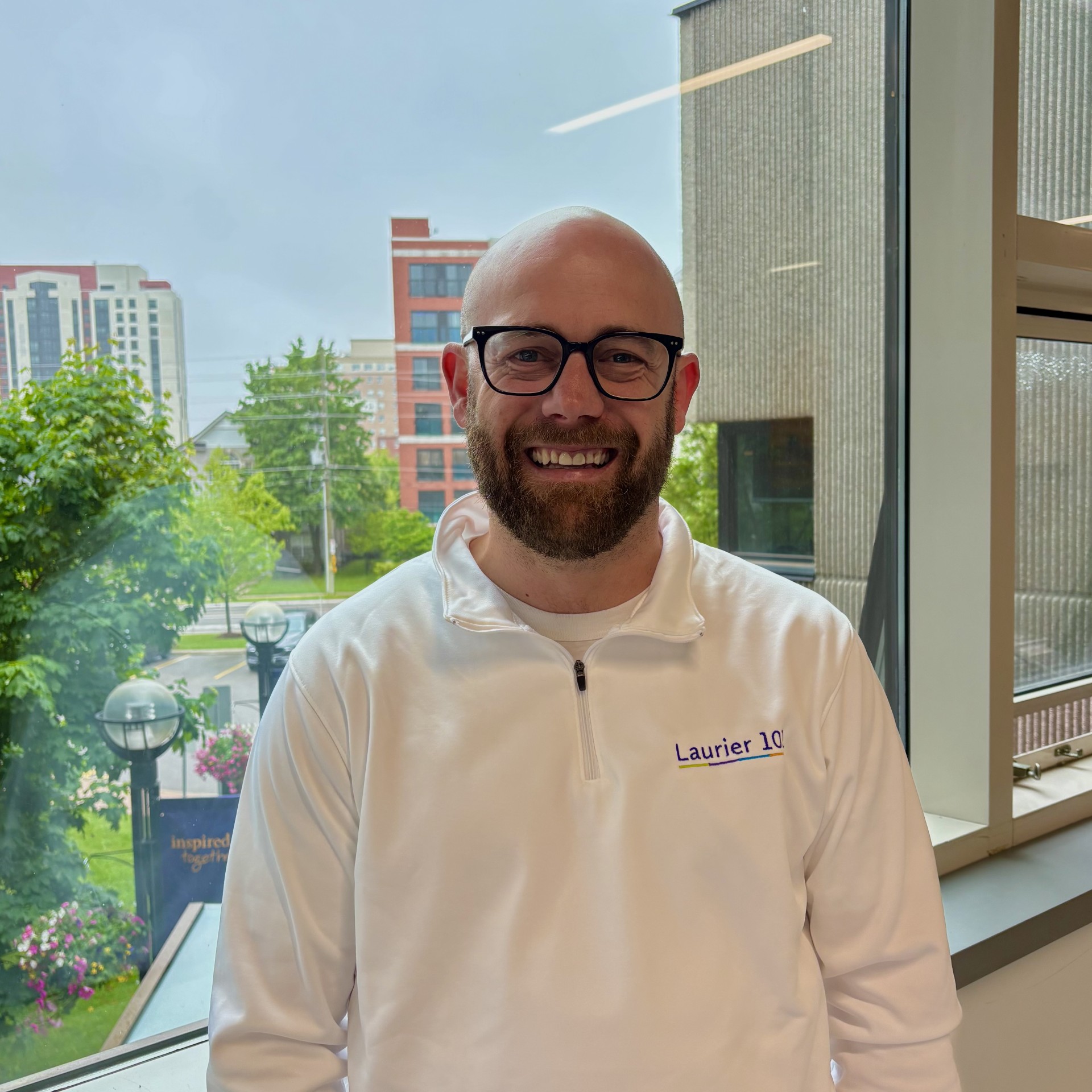 bald white man with black glasses smiling in a white Laurier shirt