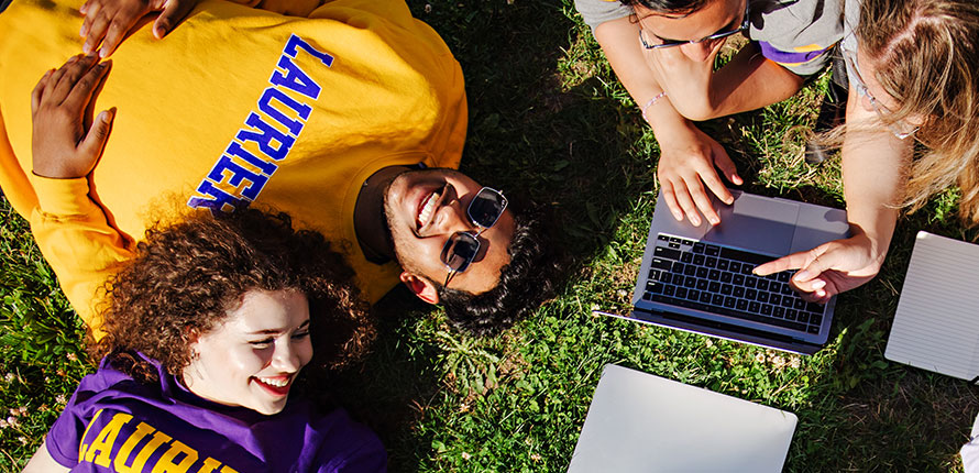 Two students lying in grass and two other students discussing homework on laptop