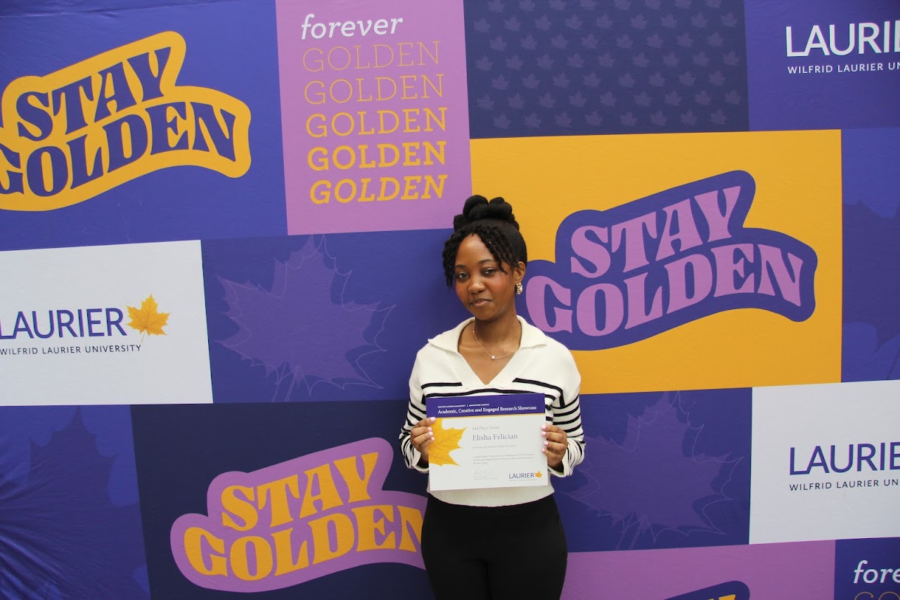 Elisha Felician holds up her award certificate in front of a colourful Laurier "Stay Golden" wall.