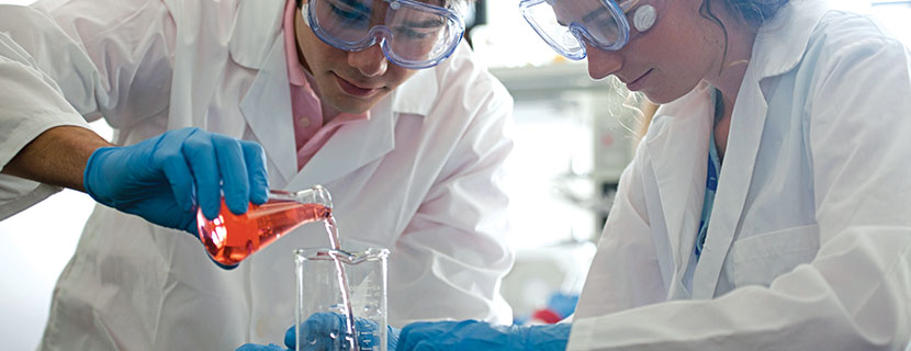 Two people in lab coats pouring liquid in a beaker.