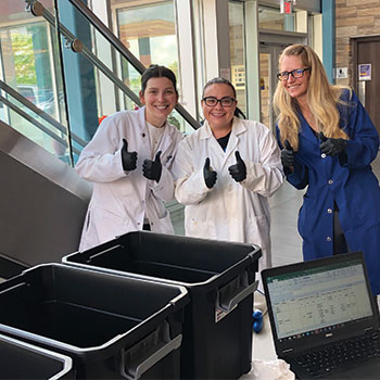 Three women in lab coats holding thumbs up beside waste bins